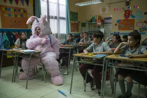 Renaldo is wearing a light pink furry outfit that looks kinda like a bunny but also has a unicorn horn for some reason. He's sitting at a desk in an elementary school classroom full of students who look bored and also confused as to why he's there in the first place.