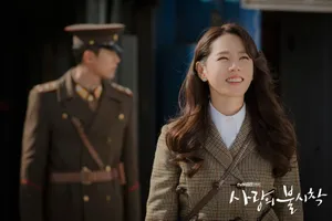 Yoon Se-ri is looking up and smiling. Her face is lit up by the sun & she's wearing a classic & stylish wool trench coat. Ri Jeong-hyeok is in the background, just out of focus, wearing his officer's uniform.