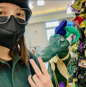 Recent selfie of me wearing a KN95 mask at the Lake Charles airport in Louisiana, standing in front of a dual Christmas and Mardi Gras display. There's Mardi Gras themed Christmas tree off to the side, where you can see part of a purple, green, and gold masquarade style mask. The tree also has a photo pinned it that appears to show a shaker of Tony Chachere's spice mix, low resolution mysterious main dish, and perhaps some cornbread on the side. But the primary focus of the photo is the aligator statue behind me. The gator is standing on its back legs at about 4 feet tall and is wearing a green and purple jester hat. You can't see much of my expression behind my mask, but trust me I was laughing. A lot.